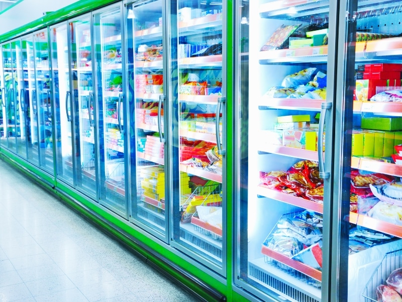 Stocked commercial freezer not cooling efficiently, displaying frozen goods behind glass doors with bright lighting and visible condensation.