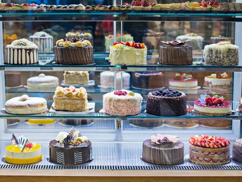 A bakery cabinet shows signs of poor cake display maintenance, with some cakes slightly askew and crumbs visible.