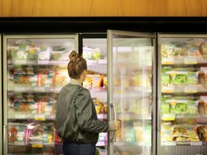A woman opens a supermarket freezer door, browsing frozen food. Learn about commercial freezer repair costs and maintenance tips.